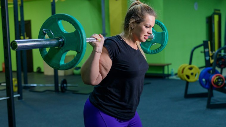 A person wearing a black t-shirt and purple leggings prepares to perform a back squat with a barbell loaded with green plates.