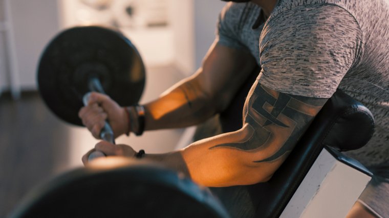 A person with a tattoo on their upper arm perfroms preacher curls with a curl bar.