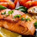 A full plate of food features cooked salmon, sliced potatoes, tomatoes, and a lemon slice.