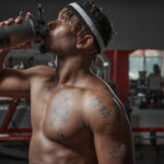 A shirtless person wearing a headband drinks from a shaker bottle in the gym.