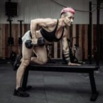 A person with short pink hair wears a sports bra while performing a single-arm dumbbell row.