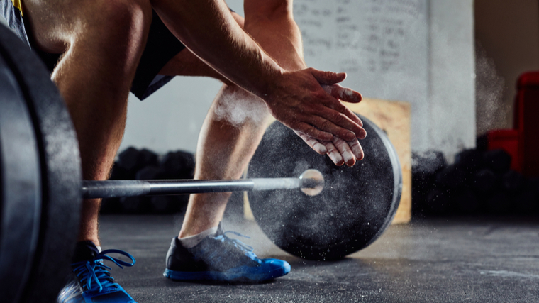 A close up image shows a persons legs, blue sneakers, and hands clapping chalk together while preparing to pull a loaded barbell off the ground.
