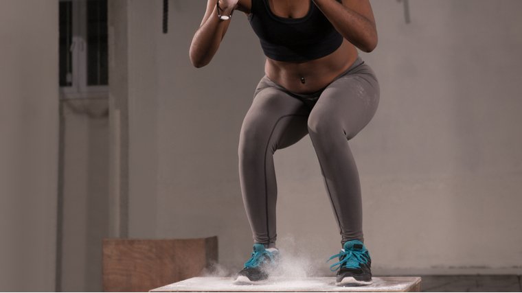 A person wearing a sports bra, grey leggings, and blue laces on their sneakers lands on a wooden plyo box as chalk sprays upward.