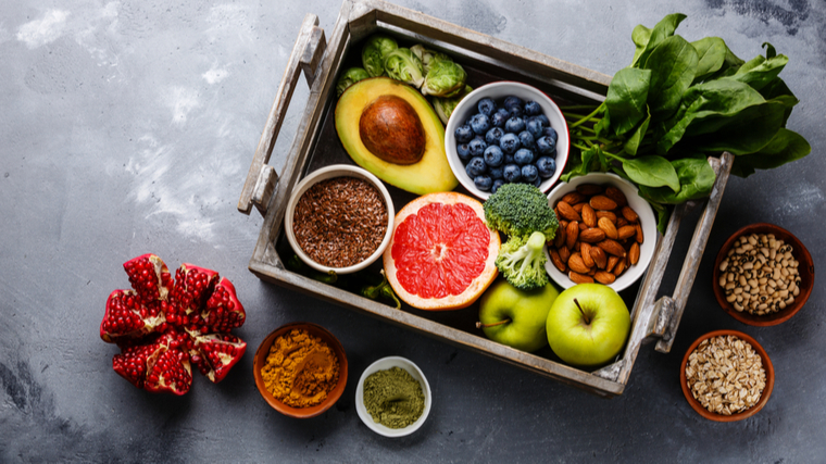 A full wooden tray of fruits, veggies, and almonds sits on a grey table alongside small bowls of seeds and a split pomegranate.