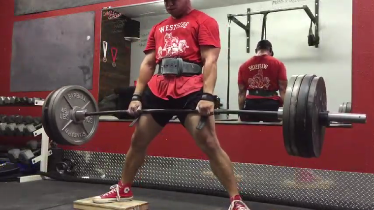 A person wearing a red Westside Barbell shirt, lifting belt, and baseball cap performs a deficit sumo deadlift.